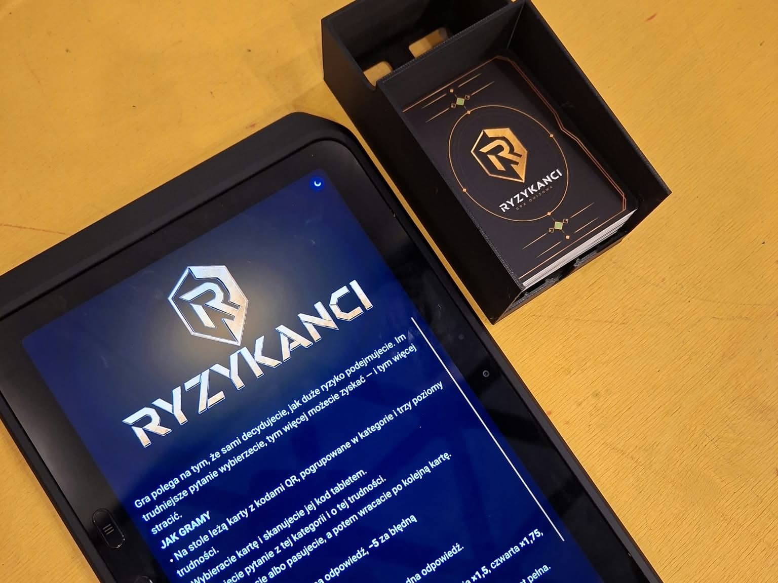Tablet z zasadami Ryzykantów i pudełko z kartami do gry na stole.