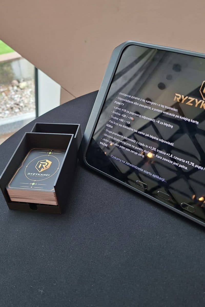 Stanowisko Ryzykantów: pudełko z kartami i tablet z zasadami gry na stoliku.