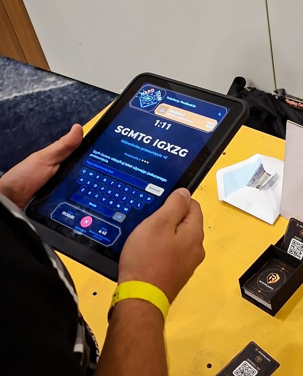 Uczestnik odszyfrowuje hasło na tablecie, obok na stole karty z kodami QR.